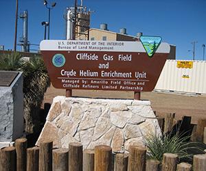 A sign reads "U.S. Department of the Interior, Bureau of Land Management, Cliffside Gas Field and Crude Helium Enrichment Unit, Managed by: Amarillo Field Office and Cliffside Refiners Limited Partnership." Behind the sign is an industrial facility with pipes, equipment, and storage containers under a clear blue sky.