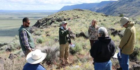 A group of six people stands in a circle on a rocky, sagebrush-covered hillside, appearing to be in discussion. Some hold papers or notebooks, and several wear hats and outdoor clothing. Behind them, a wide valley stretches out toward distant cliffs and hills under a partly cloudy sky.