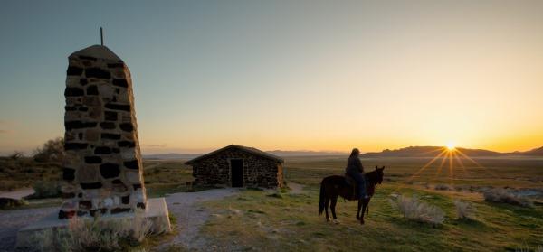 A person on a horse at sundown on a trail with a rock structure.