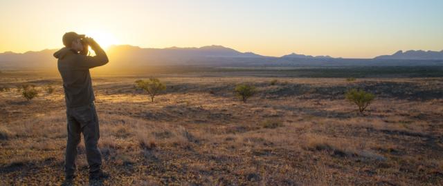 Person standing on dry grassland using binoculars to observe the distant mountains during sunset.