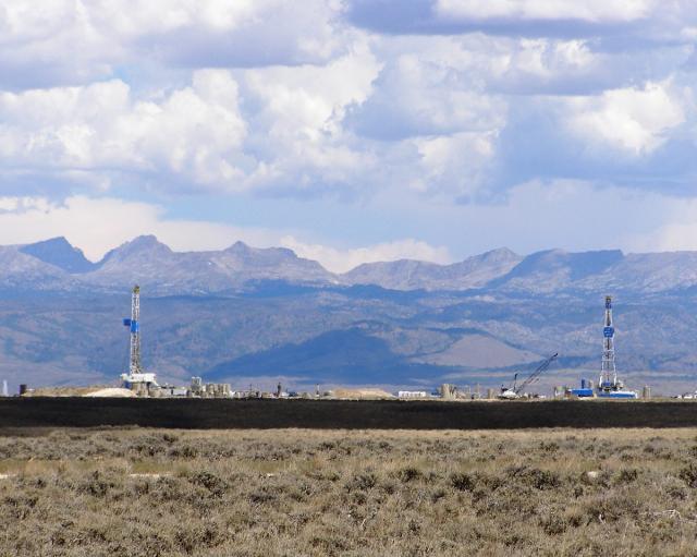 Two oil drilling rigs operate on a flat plain with rugged mountains and a cloudy sky in the background.