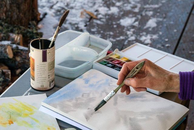 Hand holding a paintbrush applying watercolor on paper outdoors with paint palette and water containers nearby.