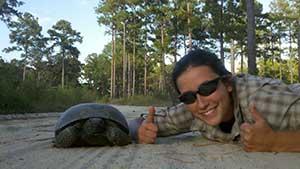 3:16 PMClaude responded: A smiling person wearing sunglasses and a plaid shirt gives two thumbs up while posing next to a tortoise on a sandy path.A smiling person wearing sunglasses and a plaid shirt gives two thumbs up while posing next to a tortoise on a sandy path. They are lying low to the ground at the tortoise's level. Behind them is a forest of tall pine trees and green undergrowth under a bright sky.