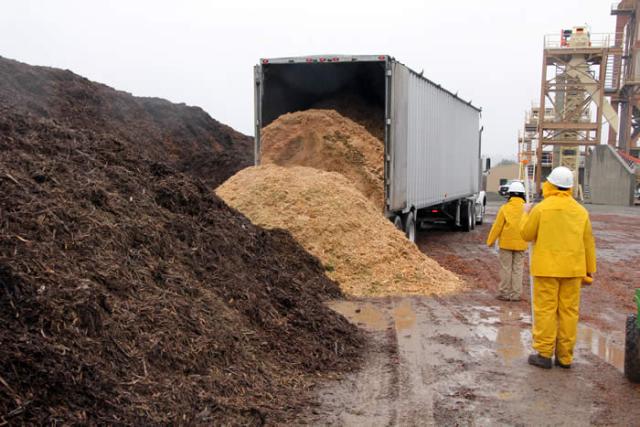 Two workers wearing yellow protective gear stand near a large truck as a load of light-colored wood chips is dumped onto the ground beside a tall pile of darker mulch at an industrial facility.