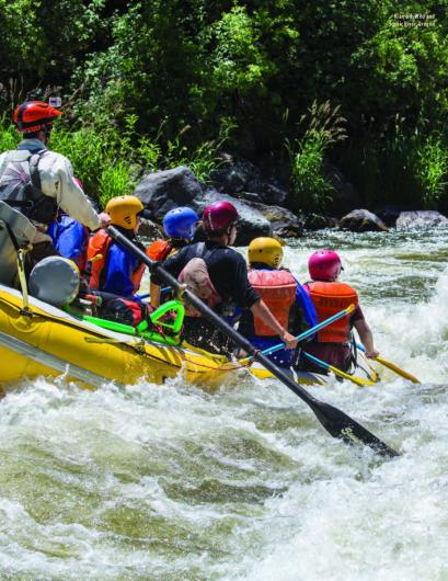 A group of people wearing helmets and life jackets paddle a yellow inflatable raft through fast‑moving whitewater, surrounded by green vegetation and rocks along the riverbank.
