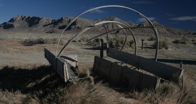 A weathered wooden wagon with broken sides and curved frame arches sits abandoned in a dry desert landscape, with rugged, eroded hills rising in the background under a deep blue sky.