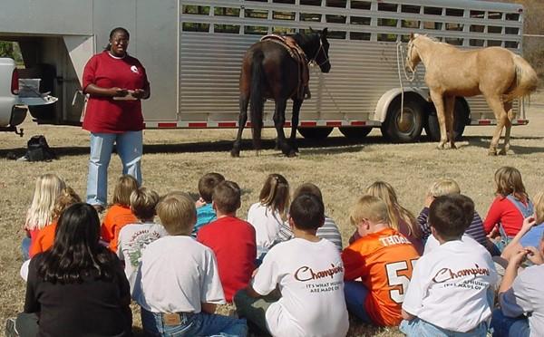 Woman teaching seated kids in front of horse trailer. 