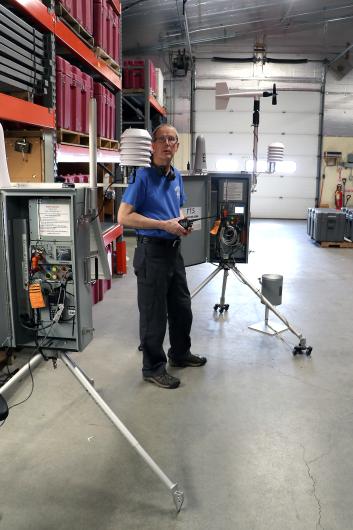 A man stands between two pieces of equipment supported by tripod legs with electronics panels and weather vanes on top.
