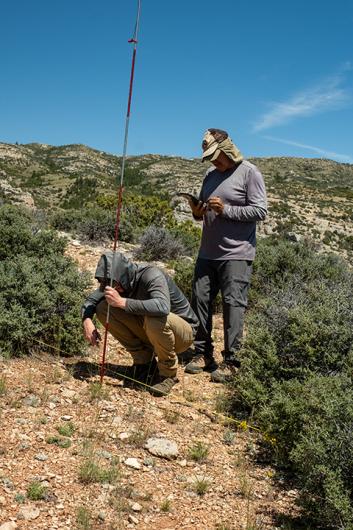 photo: one person crouches to the ground while holding pole vertically, next to another person standing and writing on a clipboard. Blue sky, brush-covered hills. 