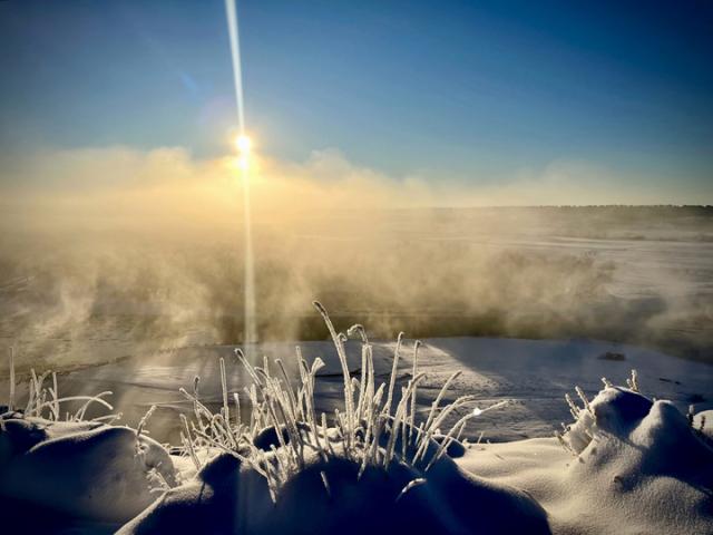 Photo. Winter snowscape of a river valley. Steam rises from the water surface. Heavy frost covers grasses sticking up from the snow-covered foreground. 