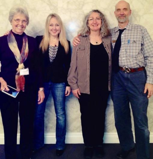 Four people—three women and one man—stand facing the camera and smiling in front of a mottled, beige-colored indoor wall.  