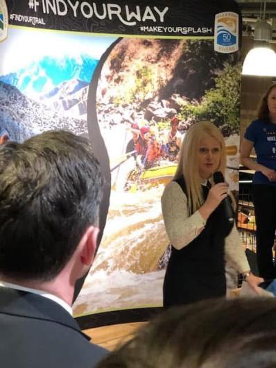 A woman holding a microphone stands in front of a display with images of snow-capped mountains and white-water rafters as she addresses members of the audience. At the top of the display are the hashtags “FINDYOURWAY,” “FINDYOURTRAIL,” and “MAKEYOURSPLASH” and a logo for Wild and Scenic Rivers.