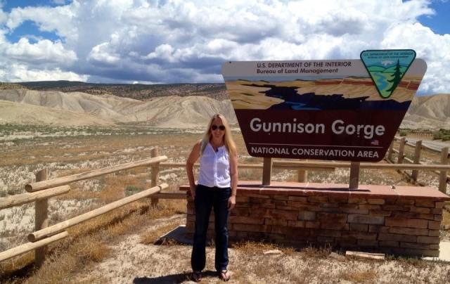 A woman in sunglasses stands facing the camera and smiling next to a sign with the BLM logo. The sign reads “Gunnison Gorge National Conservation Area.” Behind her, beyond an area of shrubland, are light brown and green mountains beneath a sky with large white clouds and patches of blue. 