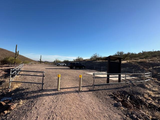 A photo showing a packed dirt path, and two bollards at the entrance to a small dirt parking lot. On the right side, near the entrance, is a concrete accessible parking spot and an informational sign.