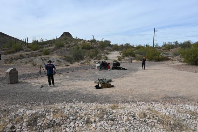 A photo showing a packed gravel shooting platform with yellow stripes separating the lanes. Two people are standing at the left lane with a tripod, two people are sitting at a table in the middle, and one person is standing on the right, observing. 