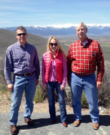 Two men and one woman stand facing the camera and smiling. One of the men and the woman are wearing sunglasses. Behind them, in the far distance, are snow-covered mountains. In the middle distance is green, hilly terrain.