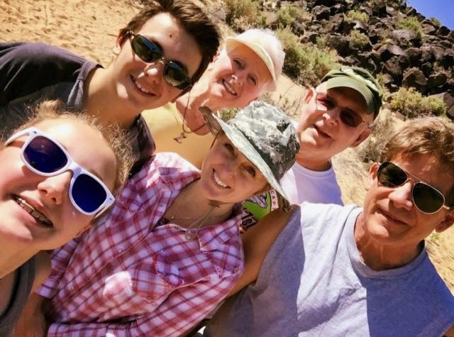In this selfie taken by her daughter, Madison, Nikki stands with her parents behind her and her husband and children on either side. The whole family is smiling, several of them wearing sunglasses and/or a hat or visor. Behind the group is brown sandy soil leading to a hilly area with large, dark rocks. 