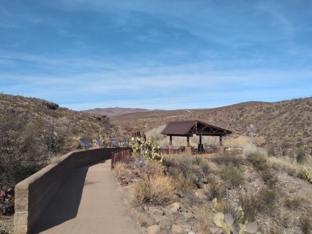 A photo showing a concrete pathway bounded by a short brick wall on the left and desert vegetation on the right. Down the path are some interpretive signs on the wall leading to a couple of benches with a shade structure overhead.