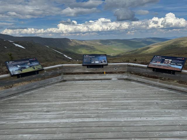 A photo showing a wooden platform with interpretive signs sitting on wooden rails around the platform, which is overlooking a gentle u-shaped valley with various shades of green on the slopes of the valley. 