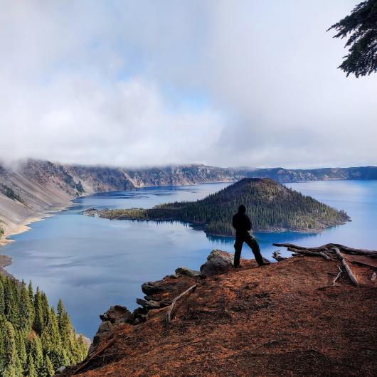 A person stands on an outcropping overlooking a mountain lake with clouds hovering over the peaks