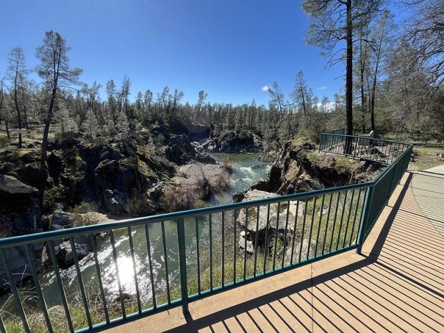 A photo showing a creek flowing in a gorge with pine trees on both sides. The foreground has a wide concrete path with a dark green metal fence for visitors to safely view the creek and canyon. 