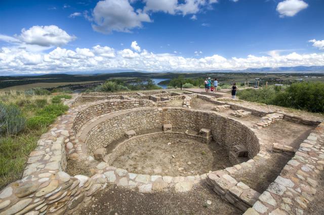A photo showing an ancient kiva, a place used for spiritual ceremonies and worship, made of stone in a circular pattern in the foreground. Surrounding the kiva are spokes of remaining stone walls that are part of the Escalante Pueblo. Behind the pueblo, an ancient indigenous settlement, you can glimpse people walking on an accessible pathway. Far in the distance is a river flowing through a green valley.  