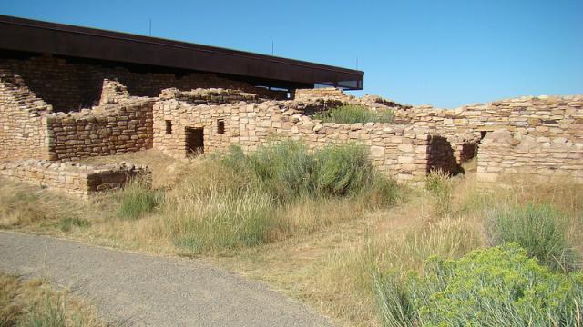 A photo showing a flat, packed gravel trail in the foreground next to the remains of a well-crafted pueblo made of sandstone blocks. The walls are about five feet high with some doorways and windows visible. A metal roof covers part of the pueblo in the background. 