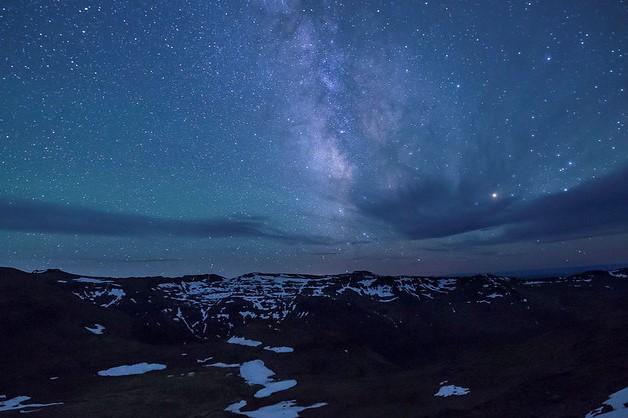 A photo showing a mostly clear night sky with some lenticular clouds on the horizon with the Milky Way as the main feature. Patches of snow are illuminated on the dark surface of hills and valleys. 