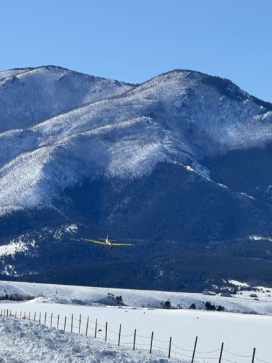 a yellow plane flies low over a snow-covered field with a mountain in the background under a blue sky
