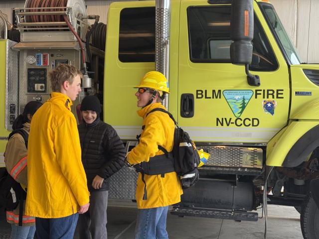 Three people in fire gear stand together and laugh in front of a yellow fire engine with BLM logo on its door.