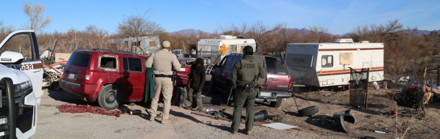 Two Rangers have their backs facing to the camera in the foreground, while several vehicles, a man, and several RVs are in the background. 
