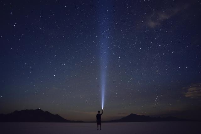 A man points a flashlight up into the sky under a starry night sky on the salt flats.