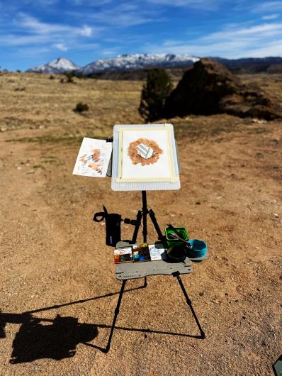 an artist's easel with a painting on it standing in a field with trees, mountains and blue skies