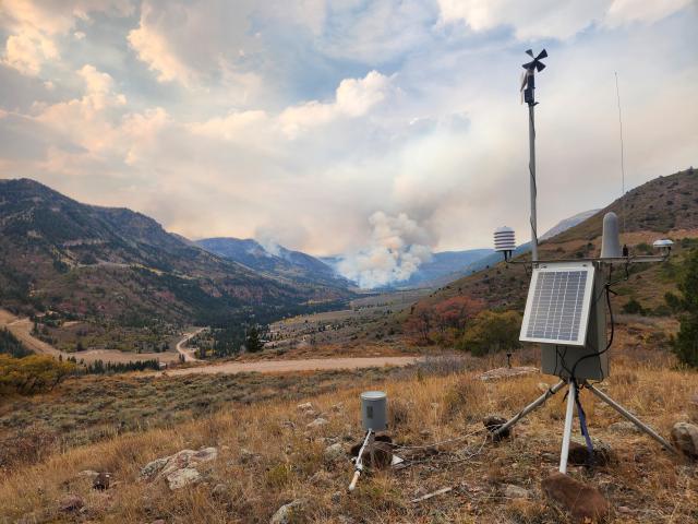 A metal structure with a solar panel and a wind vane stands on a mountain landscape with a smoke column in the distance