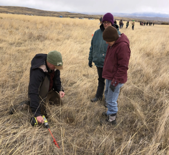 Three Caucasian people in beanies and Carhartt gear look at a hole in the ground in a brown grassy field