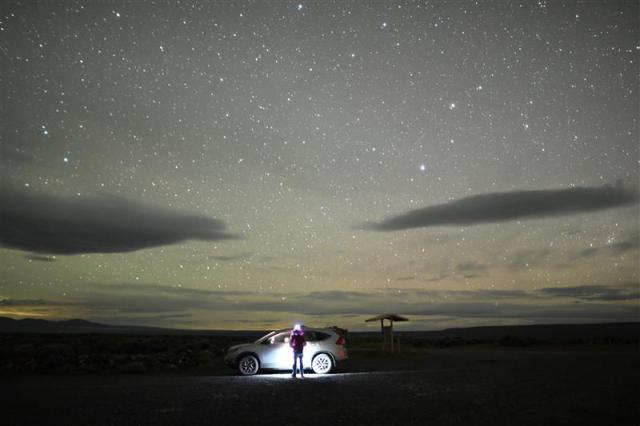 person with headlamp in front of parked car under starry sky in open empty landscape
