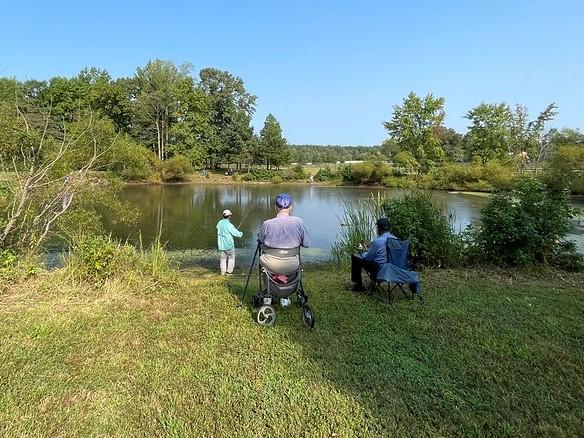 A photo showing three people on grass, fishing at a pond under blue skies. One man is standing, one is sitting in his mobility device, and the third man is sitting in a chair. The small pond has calm water. A mixed hardwood forest surrounds most of the pond in the background.  