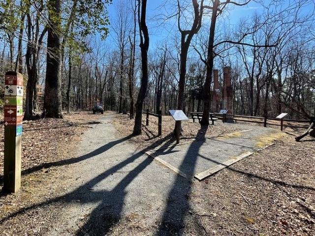 A photo showing a packed gravel pathway heading away from the camera, surrounded by trees that have dropped their leaves. The trail branches to the right towards a red brick double chimney from a former homestead. Interpretive signs and a bench are along the path near the homestead.  