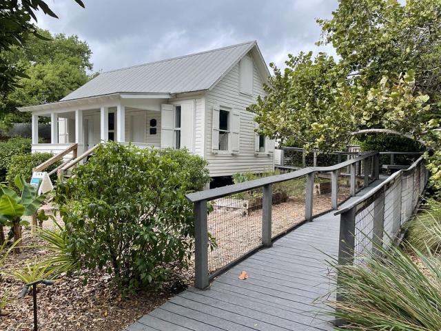 A photo showing a gray painted wood ramp with waist-high rails leading up to a white house. Shrubs and trees flank the ramp and the house. 