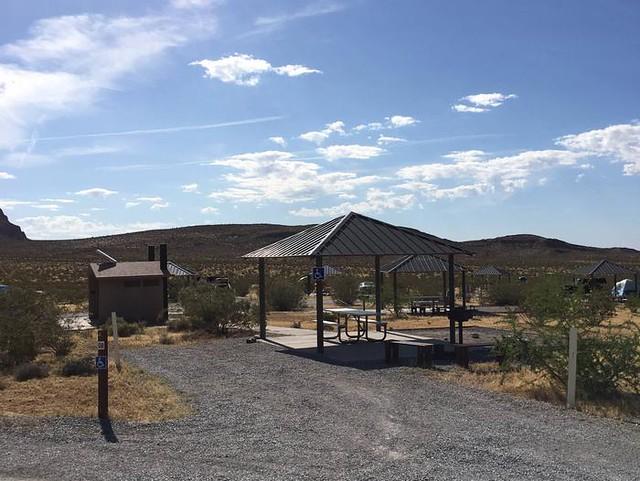 A photo showing a gravel parking spot next to a covered picnic table. A concrete path leads to a double-vault toilet, several other campsites with covered picnic tables can be seen in the background.  
