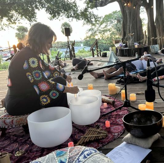 A photo showing a woman playing large white crystal singing bowls. She is dressed in black with colorful sleeves and is surrounded by candles. People can be seen laying on mats in front of her. The multi trunks of the Ficus tree are in the background. 