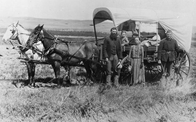 A man, woman, small child and a teenage boy stand in front of a covered wagon with two horses on a prairie..
