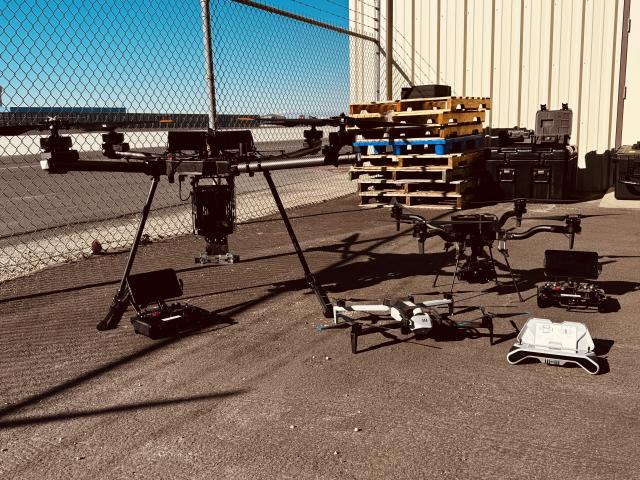 A large, medium, and small drone sit on the ground in a fence enclosed area. 
