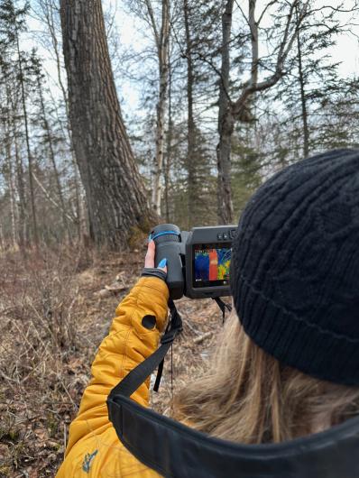 A woman is holding up a thermal imagery camera in front of a tree. 