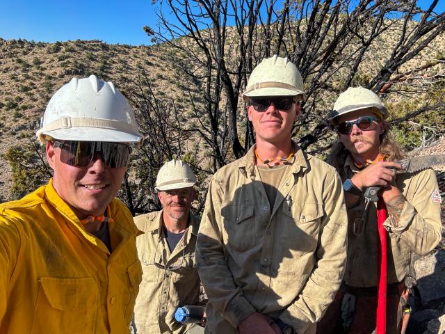 Dr. Drew stands in front of members of the Salt Lake Helitack Crew while out checking a fire. They're all wearing sunglasses, hardhats, and fire retardant shirts.