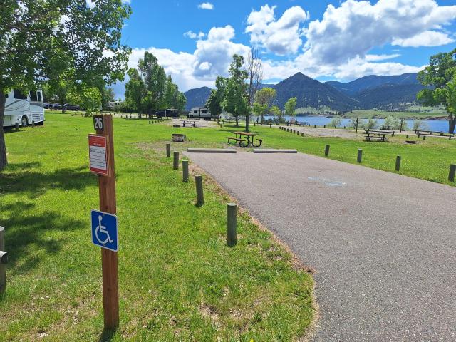 A photo showing campsite 26 with a wide paved parking pad, surrounded by trimmed grass. At the end of the parking pad is a picnic table and fire ring. Other campsites are nearby. Holter Lake and dark blue tree-covered hills can be seen in the background. 