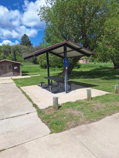 A photo showing a sidewalk leading to a concrete pad with a metal picnic table under a shelter with a cut-out on one side for a person using a wheelchair. The sidewalk continues toward an accessible double vault toilet. Trimmed grass surrounds the concrete pad and pathways. 