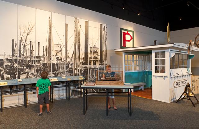 A photo showing the inside of a museum with a floor-to-ceiling, black-and-white image of steamboats, waist-high interpretive signs, a model of a boat on a table, a replica of a steamboat pilot house, and a brass bell. Achild is looking at the steamboats on the wall, while another looks at the model on the table.   