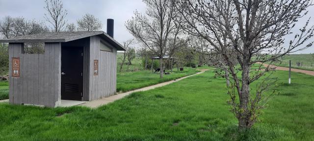 A photo showing an accessible vault toilet with a sidewalk leading to a campsite with a covered picnic table in the distance. The ground is covered with thick green grass. Leaves are just sprouting on the trees in the fore- and middle ground.  