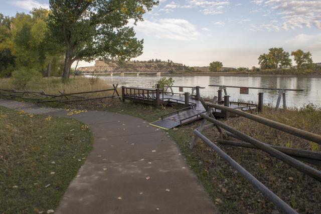 A photo showing a sidewalk curving to the left with an A-frame pole fence bordering it on the right. At the curve, a ramp connects from the sidewalk to a viewing platform on the riverbank. A short pole bumper barrier surrounds the platform for safety. In the distance a bridge crosses the river. 